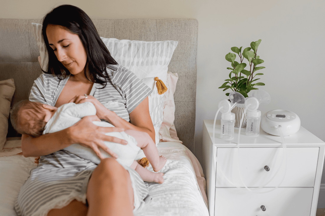 Mom is sitting on bed breastfeeding baby with cimilre breast pump on the end table