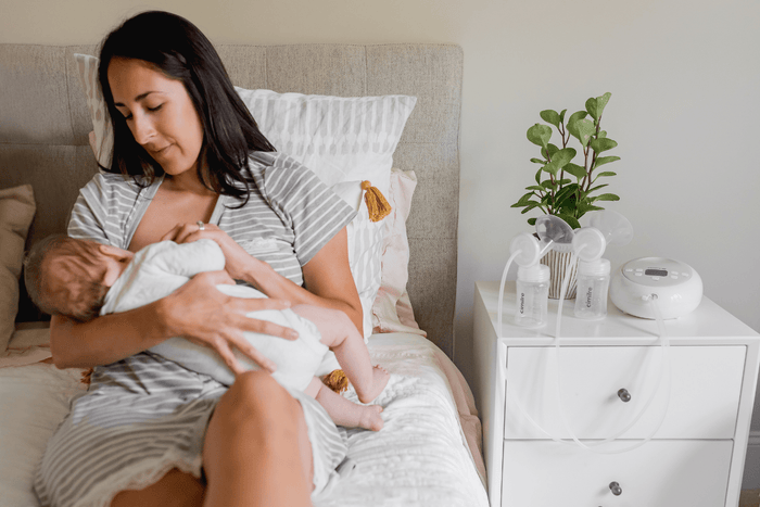 Mom is sitting on bed breastfeeding baby with cimilre breast pump on the end table
